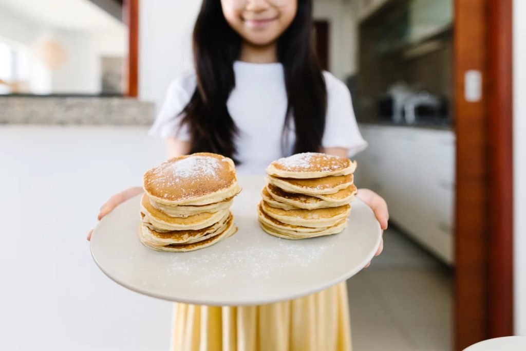a child with long dark wavy hair holding a platter with two stacks of fully pancakes