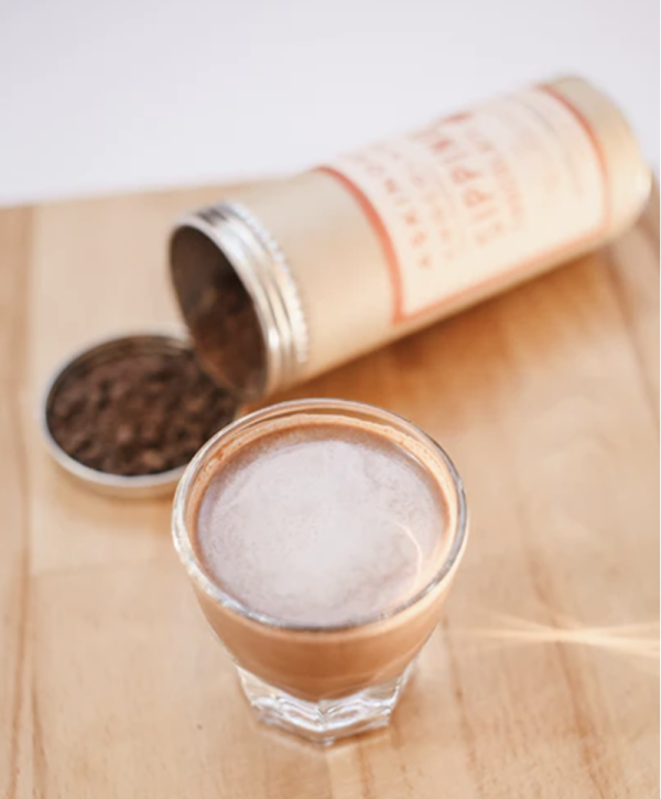 a tin of sipping chocolate in the background and a small glass filled with sipping chocolate in the foreground, all on a wooden surface